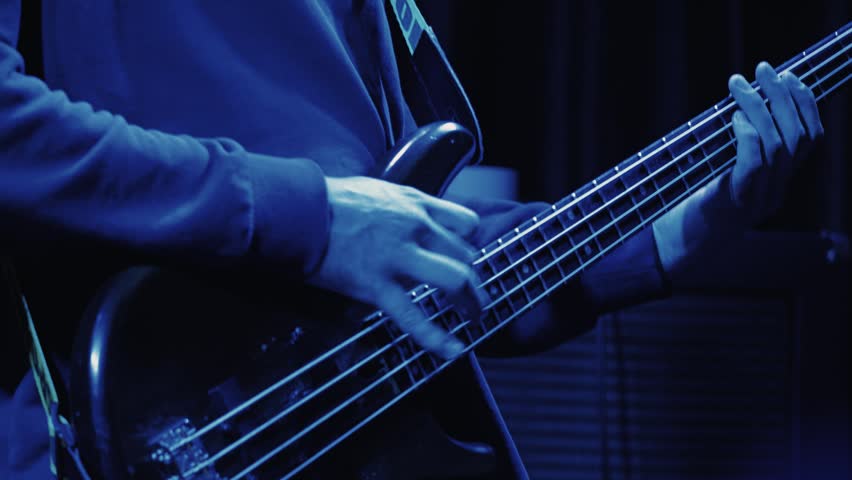 A musician plays bass guitar on stage. Close-up on hands and fretboard under dramatic purple and magenta lighting. Energetic, moody atmosphere of a rock concert or live music event