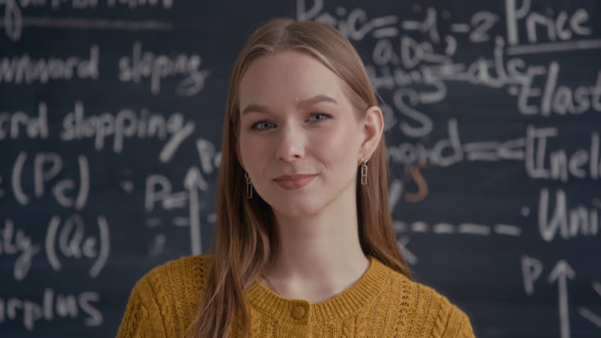 Medium close up portrait of Caucasian female student standing against chalkboard with economic notes and smiling at camera