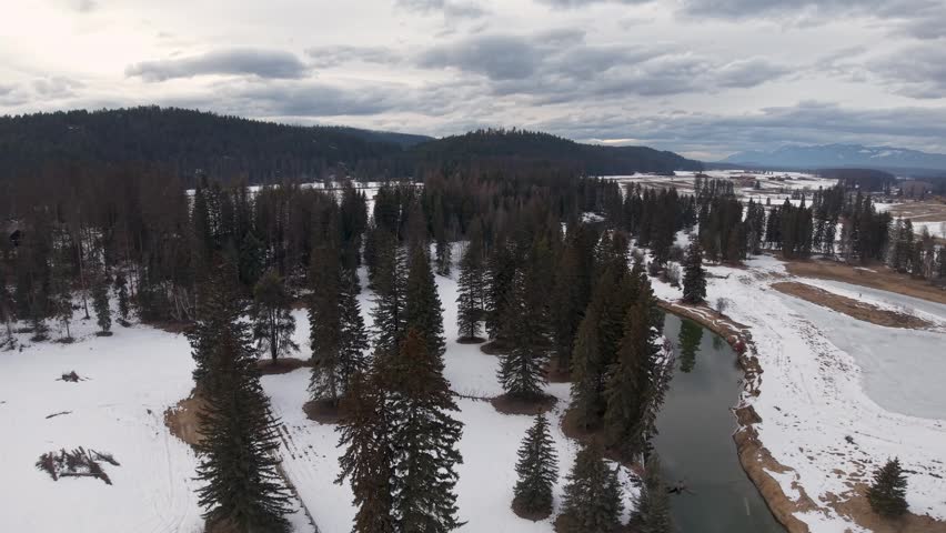 Snow covered evergreen forest and winding river viewed from above under cloudy skies with distant mountains.