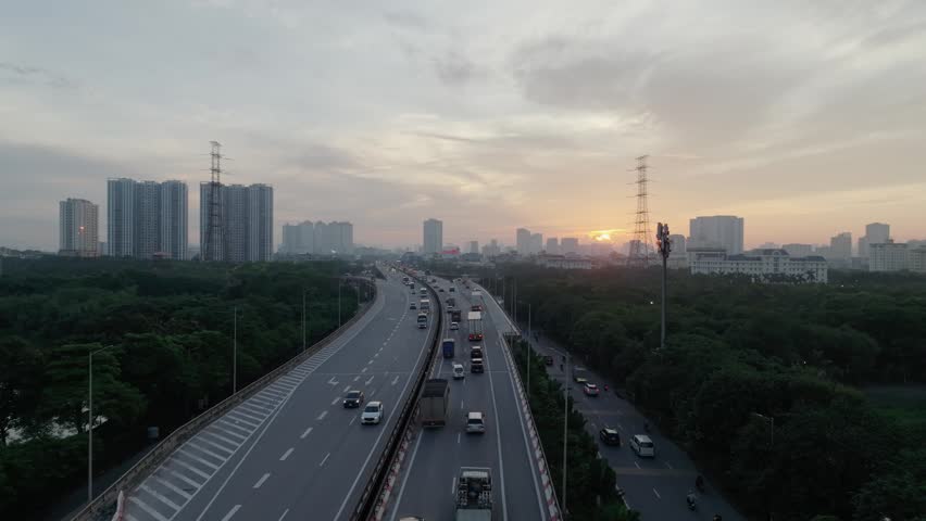 Drone perspective of multilane highway curving through green corridor in Hanoi during sunrise with vehicles moving steadily and distant high rise buildings