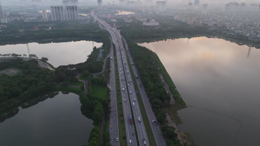 Top down aerial view of multilane highway between lakes and trees in Hanoi with steady vehicle movement and soft early morning light