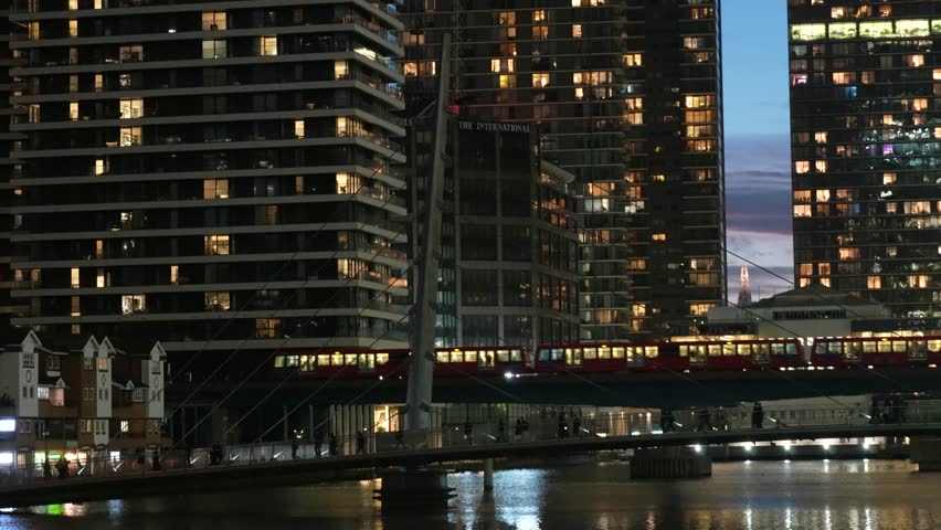 Night view in Canary Wharf, London capturing a DLR train moving across a bridge framed by illuminated skyscrapers. The scene highlights dense urban architecture, glowing windows, and reflections