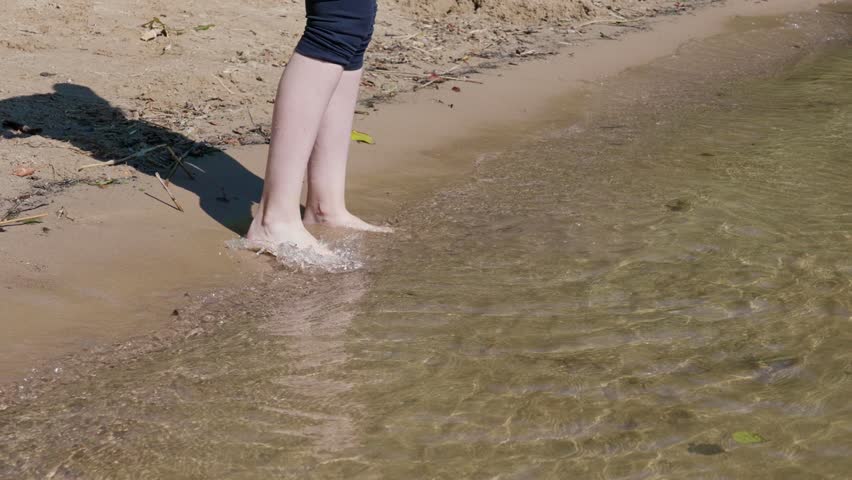 A moment of relaxation by the water, where womens legs stand barefoot on a sandy shore, entering transparent water. Leaves and seaweed are lying on the sand.