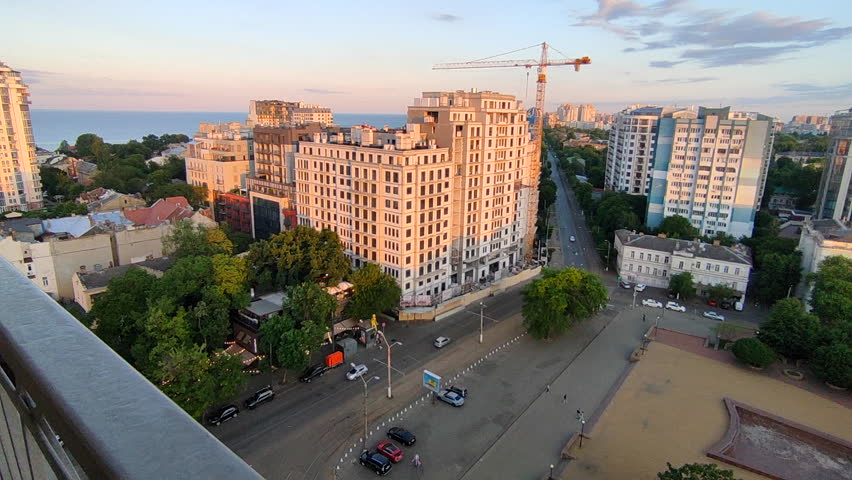 Odessa, Ukraine, August 15, 2025: City street view from above. Large multi-story buildings, square, green trees, asphalt road for cars, moving cars and walking people on sunny at sunset dawn