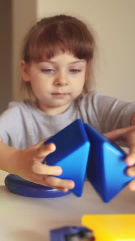 A girl is focused on arranging colorful shapes on a table. The girl interacts with blue and red shapes, showcasing her concentration and creativity in play.