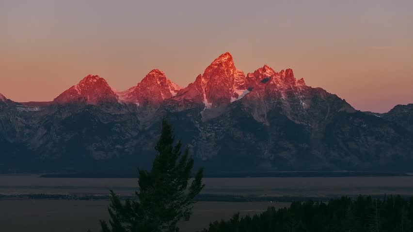 Sunrise sunset golden alpenglow on snowy Grand Teton mountain peaks above valley, aerial telephoto establishing backdrop