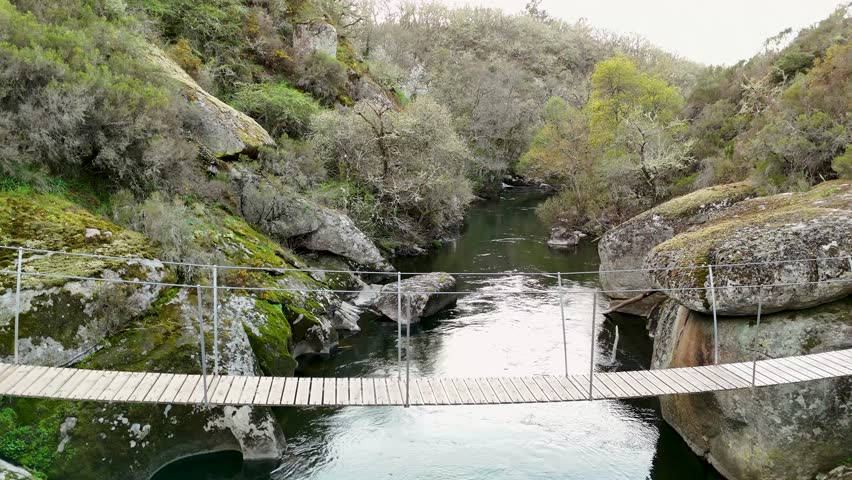 First person perspective crossing a wooden suspension bridge over a calm river in a rocky canyon