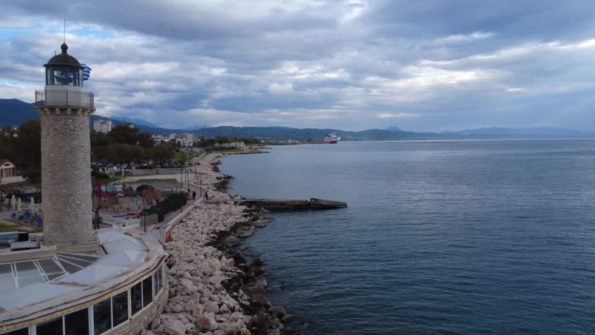 Aerial view of breakwater coastline with calm sea and cloudy sky, showing playground, coastal park with trees, lighthouse, large rocks and small pier in a seaside city in Greece, Europe.