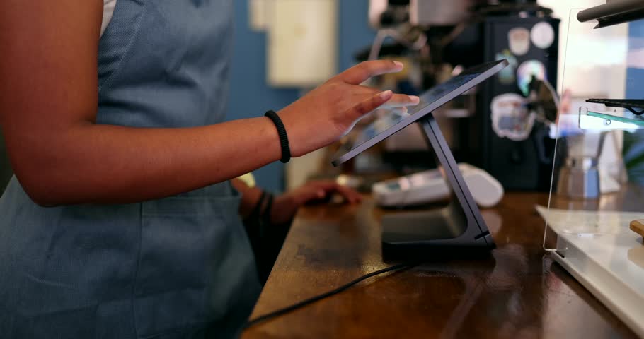 Unrecognizable african american female barista wearing an apron entering an order on a point of sale touchscreen tablet, managing transactions and serving customers in a modern coffee shop