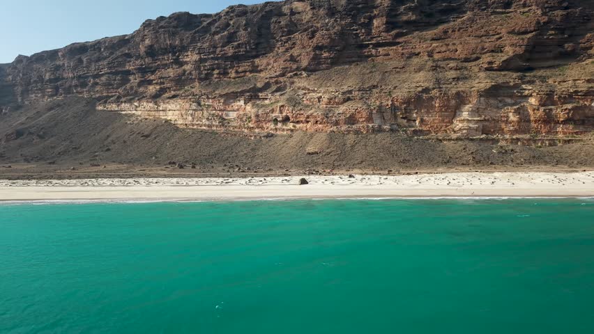 Aerial view of empty sandy beach with turquoise waves and rugged layered cliffs in a remote coastal desert landscape under clear daylight. Sokotra, Yemen.
