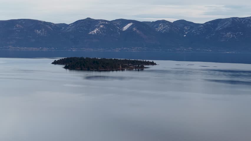Aerial view of a solitary tree-covered island in the middle of a calm lake with snow-capped mountains rising on the horizon under an overcast sky.