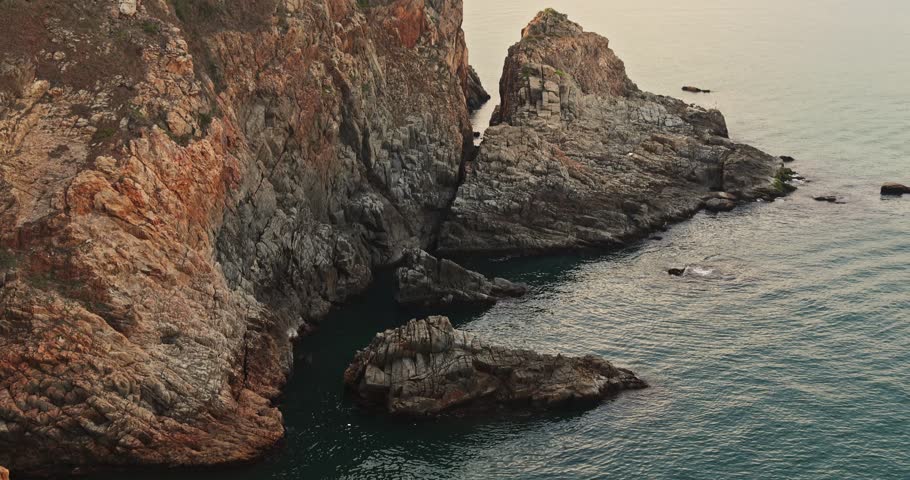 Steep rocky cliffs meet calm turquoise sea. Warm golden hour light. Rugged texture and natural coastal landscape. High-angle view of geological formations.