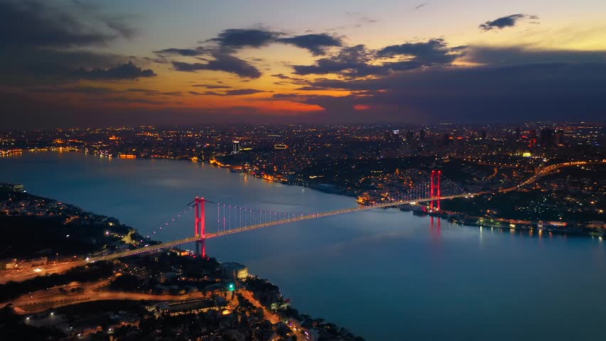 Aerial view of illuminated Bosphorus Bridge spanning Bosphorus at dusk with LED lit red towers city lights and dramatic orange sunset sky over Istanbul