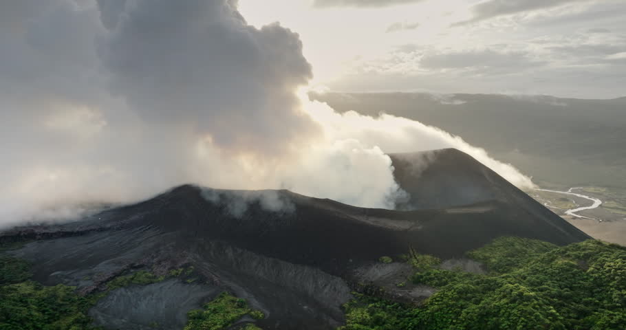 Mount Yasur volcano on Tanna Island erupting, thick white smoke and ash pouring from crater over dark volcanic slopes, framed by lush green vegetation and a river below. Aerial zoom out drone panorama