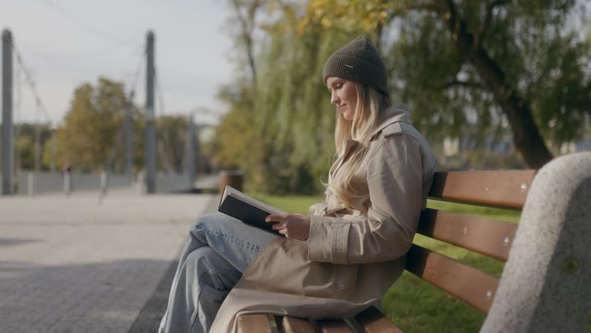 A happy young woman in a trench coat and beanie sits on a wooden park bench, laughing joyfully while reading a book during a sunny autumn day in a peaceful public park setting.