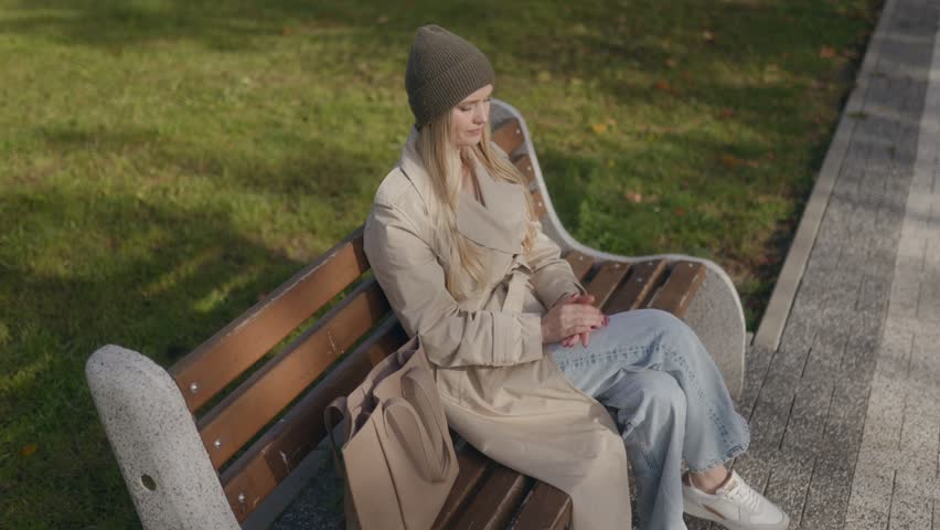 A high angle view shows a blonde woman in a trench coat sitting on a park bench, reaching into her tote bag to take out a book and start reading on a sunny autumn day.