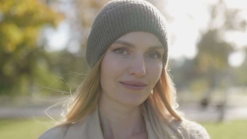 Detailed close up portrait of a beautiful smiling blonde woman in a green beanie with a soft bokeh background of golden autumn park trees under bright sunlight.