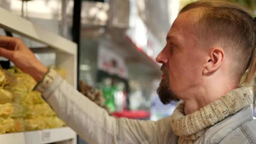Young man with stylish ponytail choosing roses at street flower shop. Man smelling red rose. Gift for beloved woman. Making surprise for wife. Celebration of women worldwide