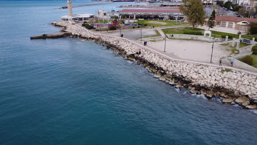 Aerial view of coastline with breakwater, harbor and calm water, showing coastal protection structure, park, lighthouse, marina, cafe, theater and people walking as waves hit rocks in Greece, Europe.