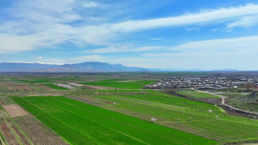 Aerial view of lush green fields stretching toward a distant town. Sky features scattered clouds over rolling hills and mountains