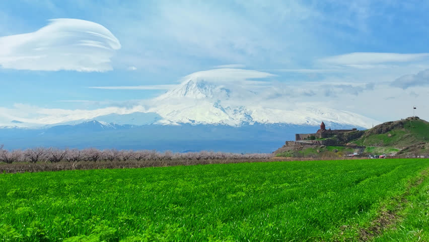 Snow-capped Mount Ararat rises majestically behind green fields. Rolling hills show blooming trees and ancient stone structures. Bright blue sky with wispy clouds enhances the peaceful scene