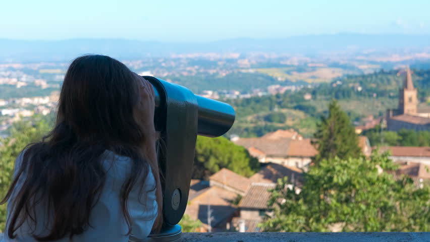 Young woman using telescope looking at cityscape. Curious young woman with long hair looking through a coin-operated telescope viewer from a high viewpoint, observing the beautiful historic cityscape