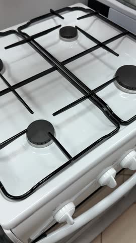 White gas stove top view closeup showing black burners and white control knobs, glossy enamel surface, subtle studio reflections.