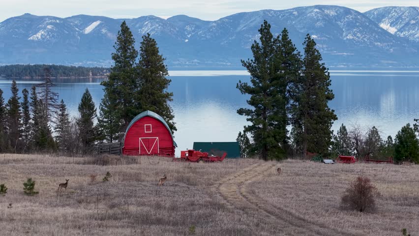 Aerial footage shows a rural landscape with a red barn on a brown field near Flathead Lake, evergreen trees, a winding path, and distant snow-capped mountains in Montana.