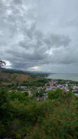 Stunning high angle view of Gorontalo city and the calm Limboto Lake from a mountain peak under a dramatic cloudy sky in Indonesia.

