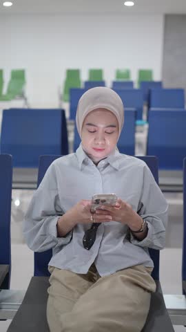 Young Muslim woman wearing hijab sitting in airport waiting area using smartphone. Vertical composition showing travel, waiting time, and modern lifestyle.