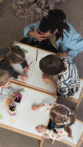 Vertical high angle shot of Caucasian children sitting around table engaged in creative kindergarten activity while drawing with colored pencils with young biracial male supervisor