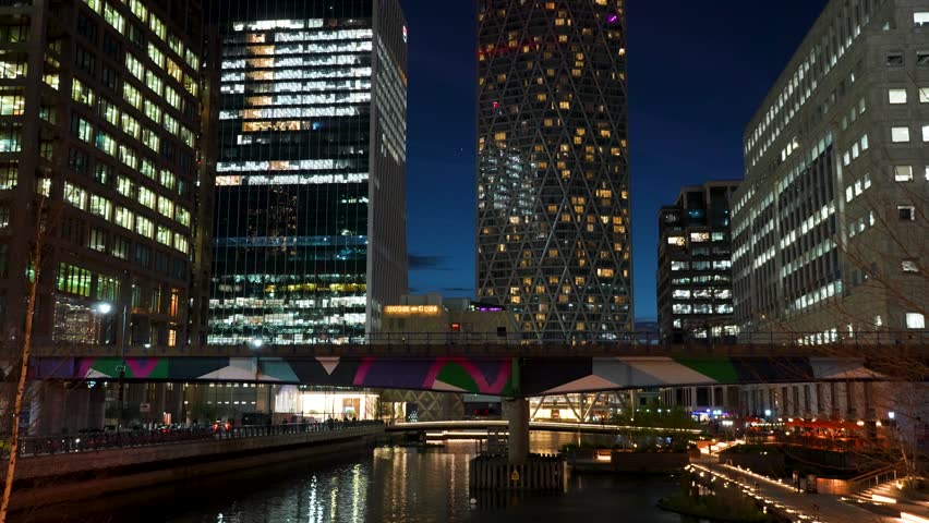 Vibrant night scene in Canary Wharf, London featuring illuminated modern skyscrapers and a metro train crossing a bridge above the water. Reflections and city lights create a dynamic urban atmosphere