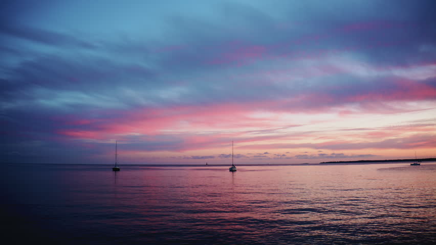 Wide shot of calm sea with sailboats anchored offshore beneath a colorful sunset sky with pink and blue clouds