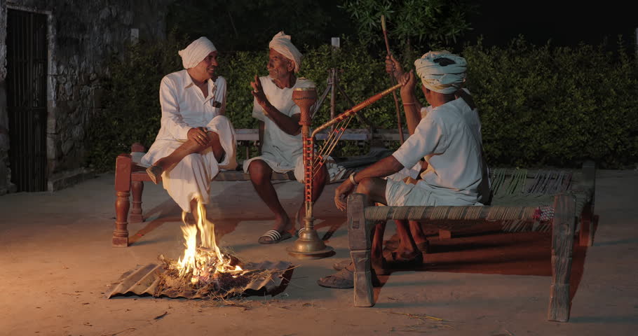 Indian old aged senior villager man wear turbans traditional white kurta pajama sitting on wooden woven cot bed near open fire. happy elderly male group talk share gossip spend time hold hookah pipe do smoking enjoy desi life outdoor night at rural area