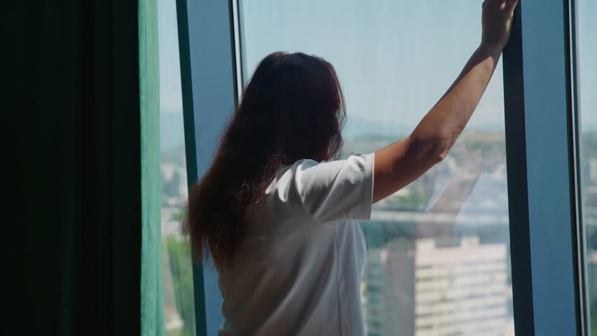 A thoughtful woman with long hair in a white shirt talks on her phone by a large window overlooking a city. She looks pensive and worried, pacing slightly during the conversation
