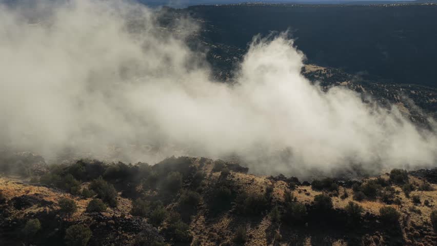 Drone shot shows thick white mist forming and rising from within a deep rugged wilderness canyon. The footage captures the atmospheric clouds moving over the rocky terrain and vegetation.