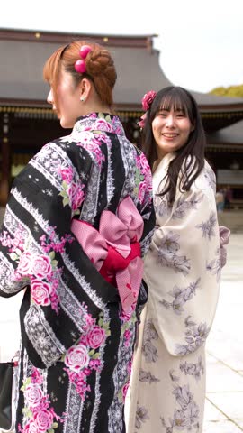 Two women wearing elegant floral kimonos turn and smile at a historic Japanese shrine courtyard