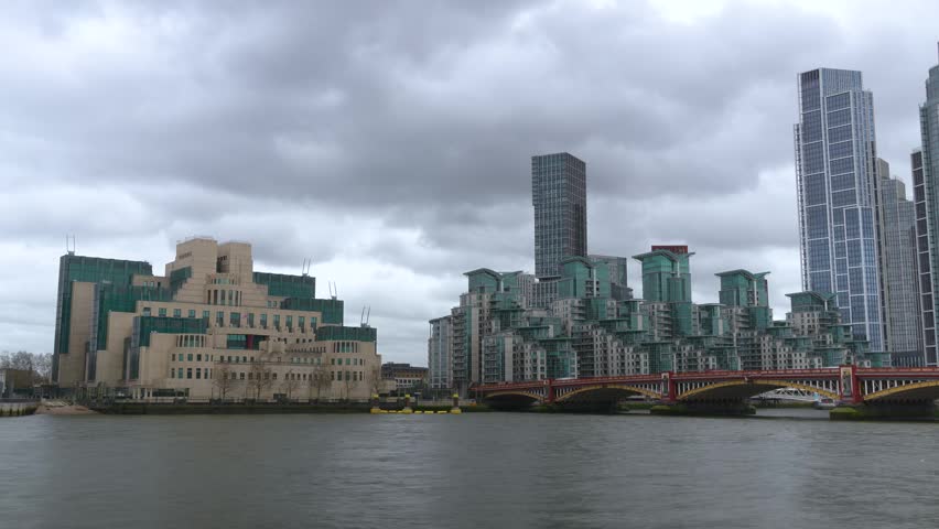 Timelapse view of the MI6 headquarters building in London alongside the River Thames, with Vauxhall Bridge and modern residential towers under dramatic cloudy skies. Iconic intelligence agency