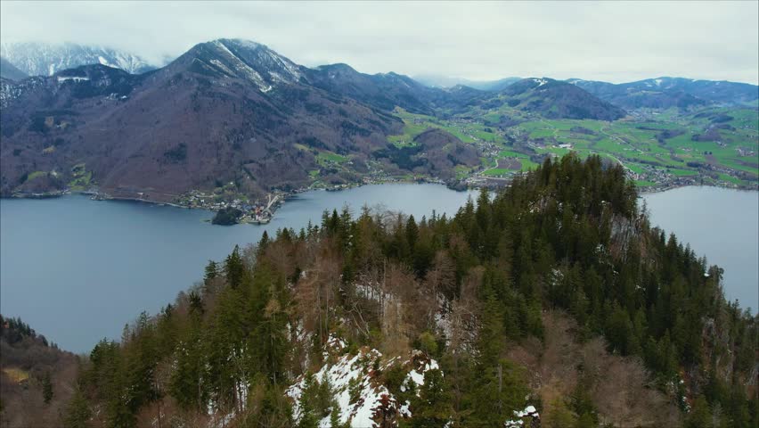Moody drone view of Traunsee, Austria, with mist drifting over snow-dusted mountains, forested slopes, and the calm blue lake beneath a heavy overcast sky.
