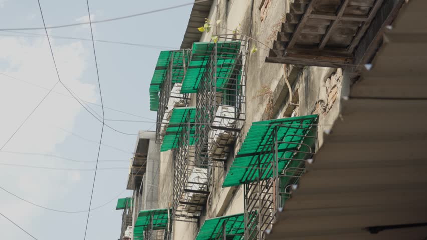 Low angle shot of a weathered residential building facade in India featuring green plastic awnings, AC outdoor unit and tangled power lines against a bright sky.