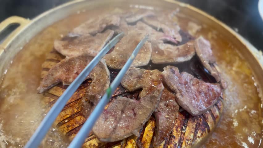 Stainless steel tongs flipping pork liver on a hot brass barbecue pan. Close-up grilling action with smoke and sizzling texture, showing cooking process in restaurant style.