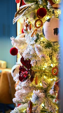 Hands of woman decorating Christmas tree with ornaments and berries. Close up of female hands hanging decorations on snow covered Christmas tree with warm lights and bokeh background.