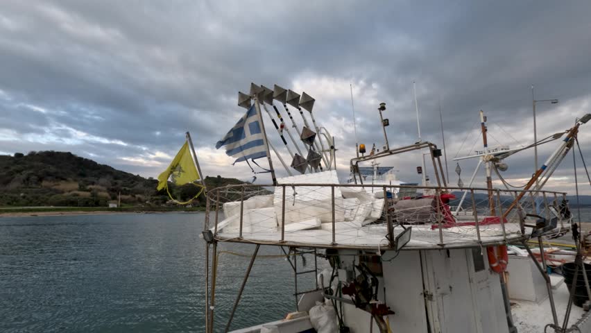 A fishing boat sits in the harbor as windmill sails turn in the breeze. The boat is surrounded by water with hills in the background. The sky is cloudy and gray.