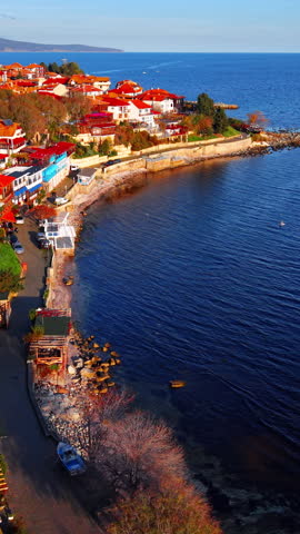 Seaside town featuring traditional architecture with red roofs, blue sea water, and a stone embankment. Aerial view of coastal town. Vertical video.
