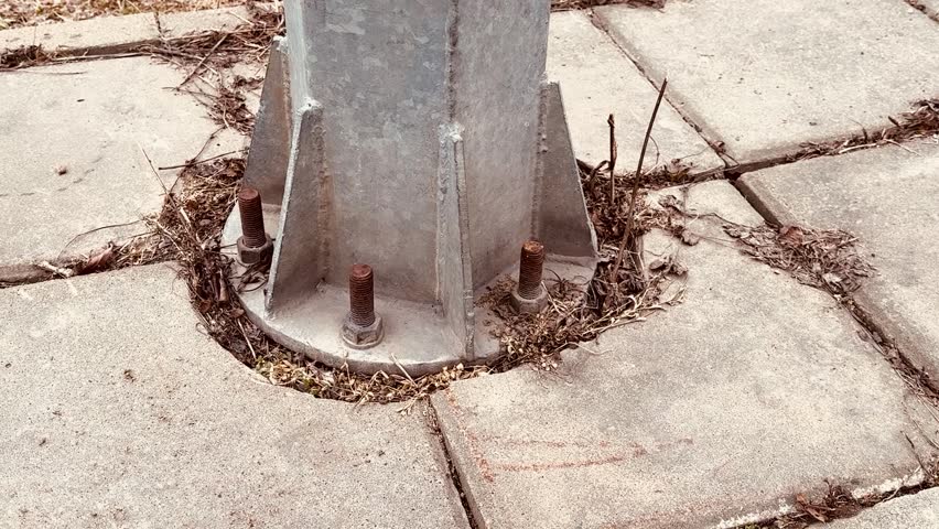 Rusted sign pole base on concrete, closeup of anchor plate bolted to sidewalk with corroded bolts, dried weeds and moss.