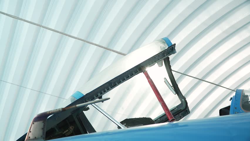 Raised glass roof of fighter pilot cockpit in sunlight, close up. Open cockpit of blue airplane standing in hangar without people.