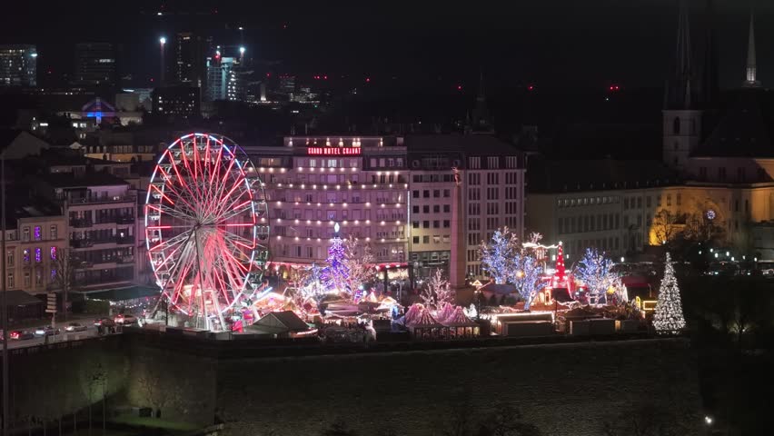 Night drone footage of Monument du Souvenir in Luxembourg with illuminated Christmas market and Ferris wheel, capturing festive lights, holiday atmosphere, and vibrant winter city scene.
