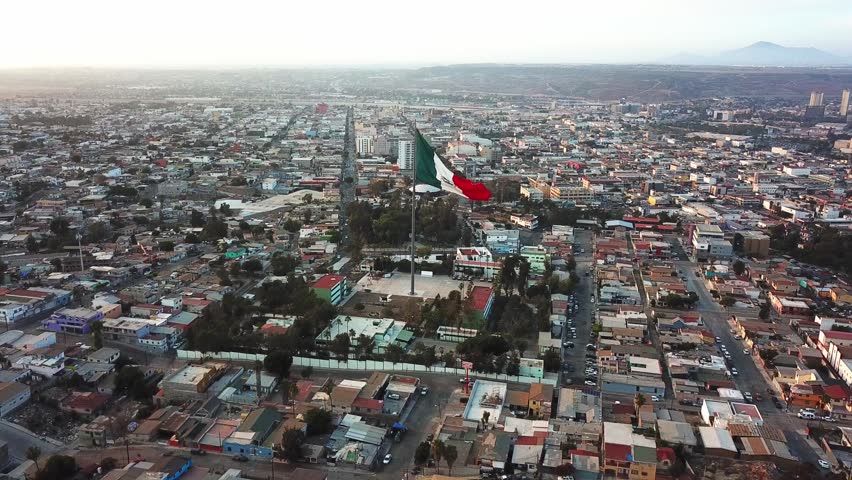 Drone shot of a large Mexican flag waving over an urban area in Tijuana, Baja California, surrounded by dense city streets and neighborhoods.