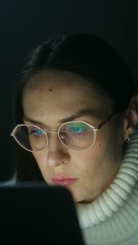 Focused young woman wearing glasses working late at night on a laptop, with the screen light reflecting in her eyewear while she stares intently at the monitor in a dark room. Vertical