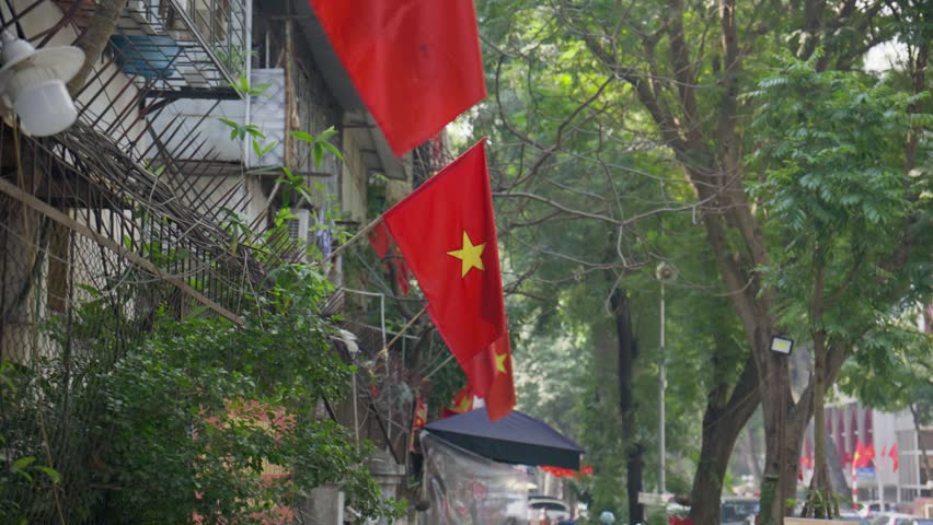 Vietnamese flags waving in wind on building balcony with lush trees and urban traffic in background on typical day in city of Hanoi, Vietnam